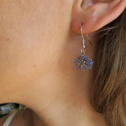 A close-up of a person's ear wearing a dangling flower earring. The earring has a silver-toned fishhook back and a small purple flower charm with a yellow center, shown against skin and brown hair.