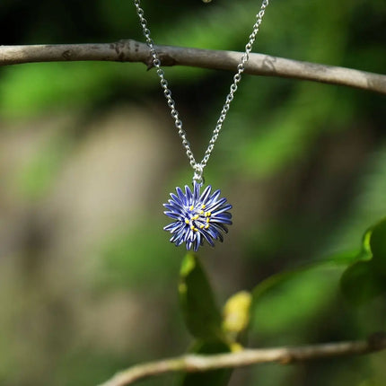 A silver chain necklace with a flower-shaped pendant featuring blue petals and a yellow center, hanging from a tree branch against a blurred green natural background.