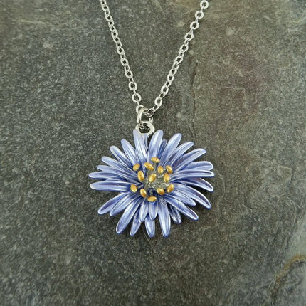 A silver chain necklace with a daisy flower pendant, featuring light blue petals and a gold center, displayed on a textured gray stone background.