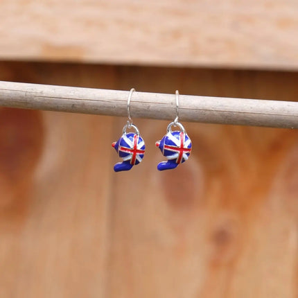 A pair of silver-toned dangle earrings with small enamel bear charms decorated in a red, white, and blue Union Jack pattern, hanging from a horizontal wooden rod against a warm, blurred wood background.