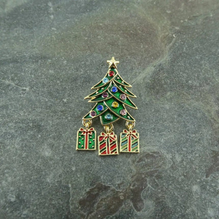 A gold-tone Christmas tree brooch with green enamel leaves, multicolor rhinestones, and three dangling gift boxes, displayed on a gray stone surface.