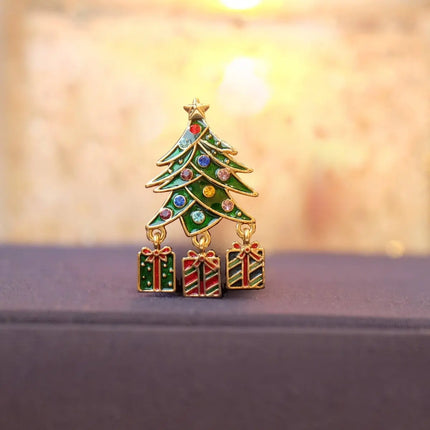 A Christmas tree and gift box enamel pin with gold-tone metal trim and colorful rhinestones, displayed on a dark surface with a warm, blurred background.