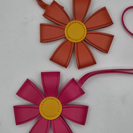 Two flower-shaped leather bag charms or pouches on a light gray background. The top one is terracotta orange with a yellow center, and the bottom one is magenta pink with a yellow center.