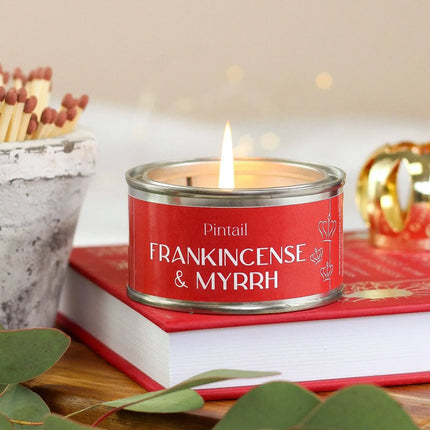 A red scented candle tin labeled 'Pintail FRANKINCENSE & MYRRH' sits on top of a red book, with a lit flame, decorative matches, green leaves, and a softly blurred warm background.