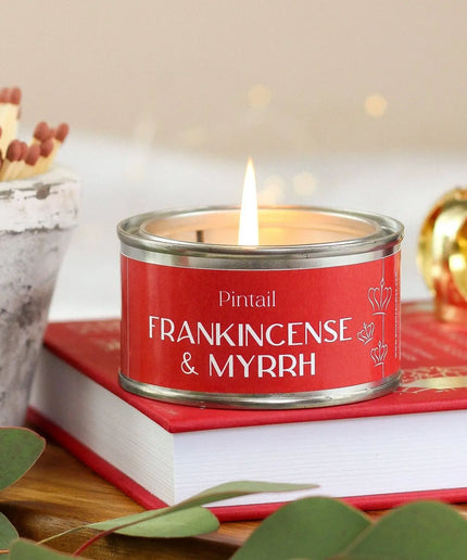 A red scented candle tin labeled 'Pintail FRANKINCENSE & MYRRH' sits on top of a red book, with a lit flame, decorative matches, green leaves, and a softly blurred warm background.