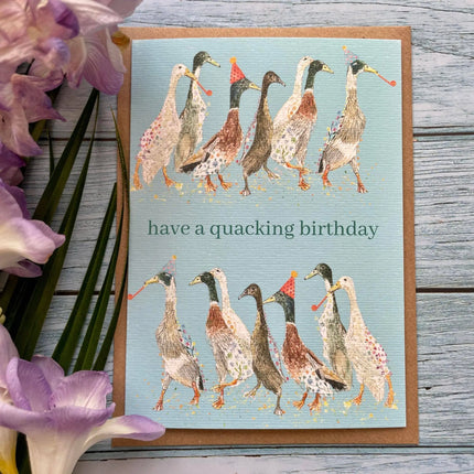 Greeting card with ducks illustration and 'have a quacking birthday' text on a wooden surface with flowers.