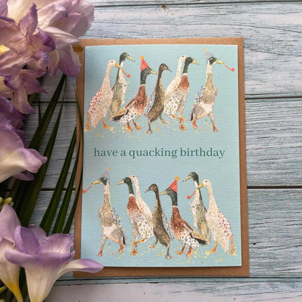 Greeting card with ducks illustration and 'have a quacking birthday' text on a wooden surface with flowers.
