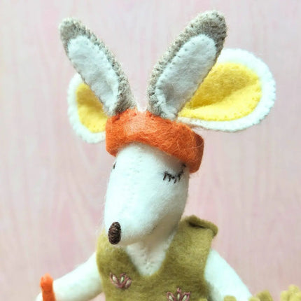 A close-up of a handmade felt animal toy, likely a rabbit or kangaroo, with long white and gray ears, yellow inner ears, an orange headband, and a green vest, set against a soft pink background.