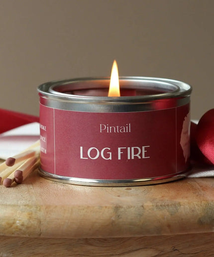 Candle in a red tin labeled 'Log Fire' by Pintail, with matches on a wooden surface.