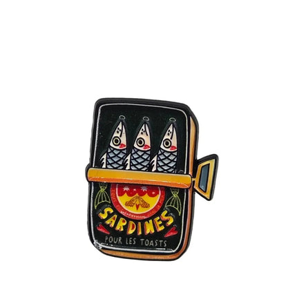A small enamel pin shaped like a sardine tin, featuring three sardines on a black background with yellow, orange, red, and white details, and the text 'SARDINES FOUR LES TOASTS' on a light gray surface.