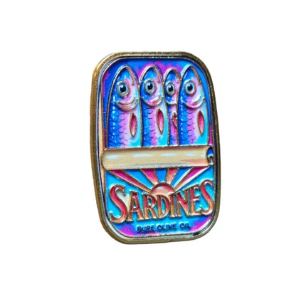 A vintage Sardines enamel pin with a gold-plated border, featuring three sardines and the Sardines logo in vibrant blue, pink, and purple colors, resting on a wooden surface.