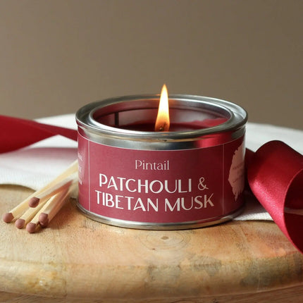 Candle in a red tin labeled 'Pintail Patchouli & Tibetan Musk' with matches and a red ribbon on a wooden surface.