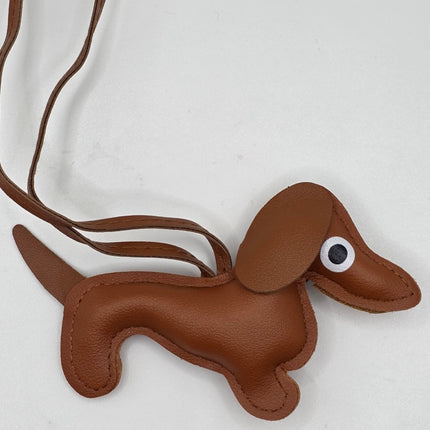 A brown faux-leather dachshund-shaped bag charm or crossbody bag with a long strap, featuring a white and black plastic eye on its side, shown on a light gray background.