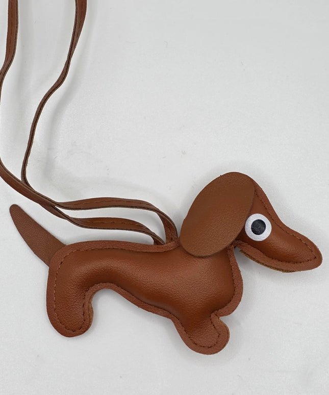 A brown faux-leather dachshund-shaped bag charm or crossbody bag with a long strap, featuring a white and black plastic eye on its side, shown on a light gray background.