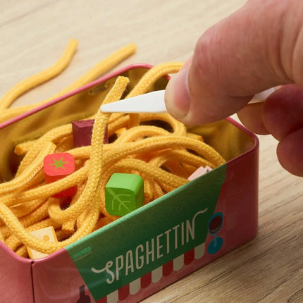 A close-up of a hand using a white plastic pick to remove yellow spaghetti-shaped cords from a pink and green tin labeled 'SPAGHETTIN', with small colorful cube-shaped pieces visible inside.